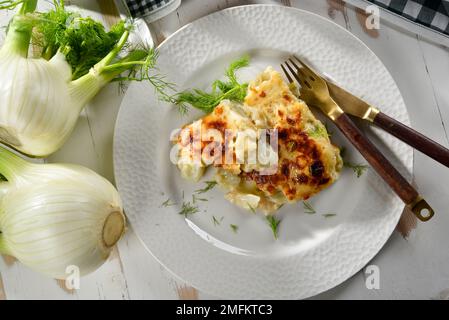 Finocchio al gratin al forno servito in un piatto bianco, con finocchio fresco nelle vicinanze Foto Stock