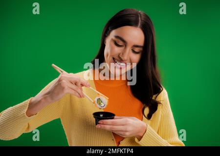 donna sorridente brunette che tiene sushi roll vicino salsa ciotola isolato sul verde, immagine stock Foto Stock