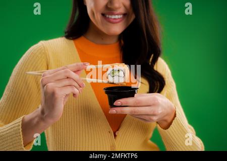 vista ritagliata di una donna sorridente che tiene sushi roll vicino salsa ciotola isolato sul verde, immagine stock Foto Stock