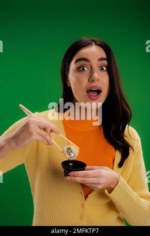 donna stupita che tiene salsa ciotola e bacchette con sushi roll mentre guarda la fotocamera isolato su verde, immagine stock Foto Stock