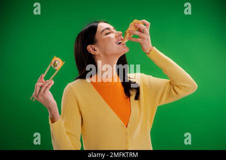 giovane donna bruna con bastoncini di pollo con sushi roll mentre mangiare pizza isolato sul verde, immagine stock Foto Stock