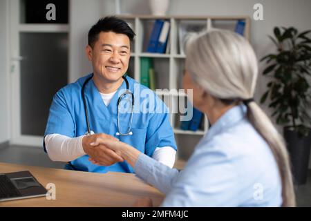 Allegro medico coreano di mezza età che scuote le mani con vecchia donna caucasica paziente in clinica Foto Stock