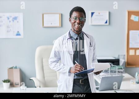 Vita in su ritratto del giovane medico afro-americano in piedi dalla scrivania in clinica e tenendo appunti sorridendo alla macchina fotografica Foto Stock