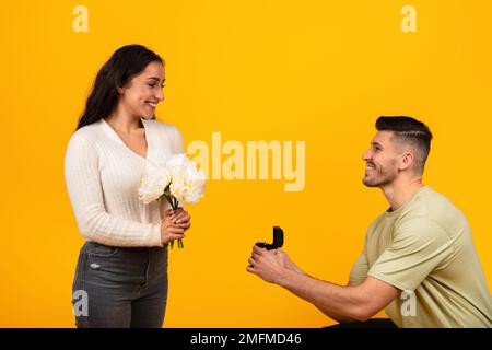 Giovane ragazzo arabo allegro in casual sul ginocchio dà scatola con anello a donna con fiori Foto Stock