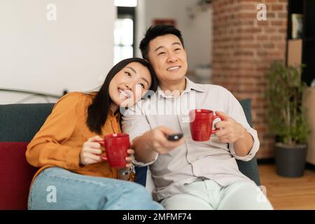 Felice coppia asiatica sposata che passa il tempo a casa insieme, bere caffè e guardare la tv, rilassarsi sul divano Foto Stock