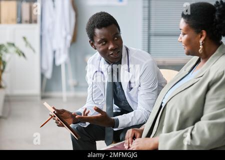 Ritratto di giovane uomo nero come medico di consulenza femminile paziente in clinica e tenendo tablet in sala d'attesa Foto Stock