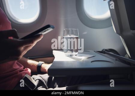 Bicchiere d'acqua sul tavolo del sedile in aereo. Il passeggero utilizza il telefono durante il volo. Foto Stock