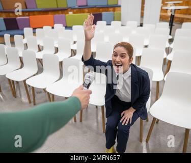 Una donna d'affari caucasica dai capelli rossi siede in prima fila in una sala conferenze vuota e risponde in un microfono allungato. Foto Stock