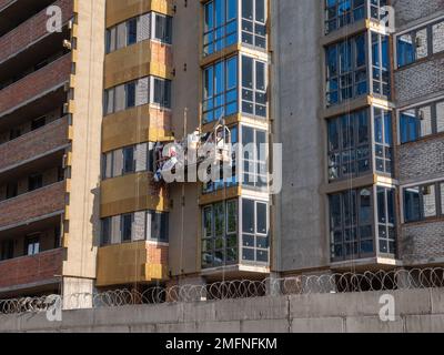 Costruttore maschile in una costruzione sospesa culla lavori su una facciata di un alto edificio di recente costruzione. Nuovo distretto in costruzione a Kiev. Foto Stock