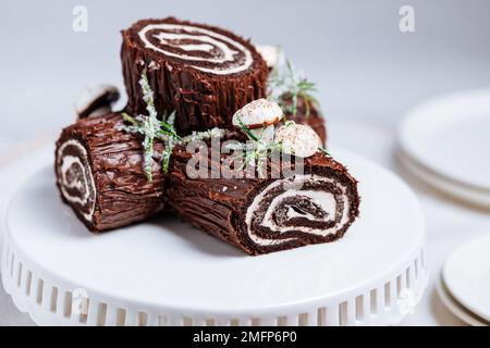 Dessert francese chiamato Yule log o bûche de Noël con funghi merengue e foglie in cima alla vetrata al cioccolato. Posto di fronte all'albero di Natale. Dic Foto Stock