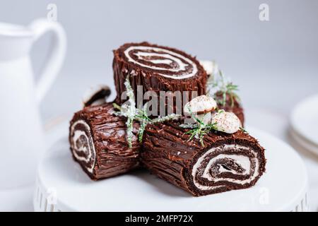 Dessert francese chiamato Yule log o bûche de Noël con funghi merengue e foglie in cima alla vetrata al cioccolato. Posto di fronte all'albero di Natale. Dic Foto Stock