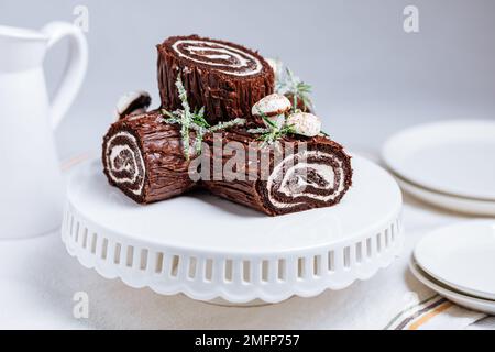 Dessert francese chiamato Yule log o bûche de Noël con funghi merengue e foglie in cima alla vetrata al cioccolato. Posto di fronte all'albero di Natale. Dic Foto Stock