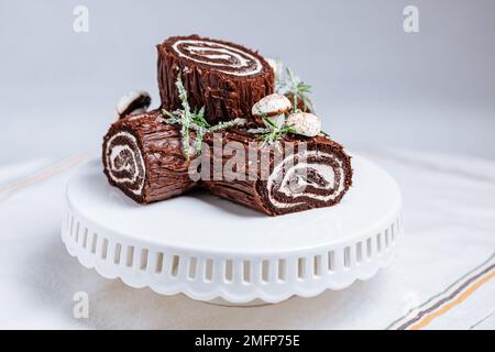 Dessert francese chiamato Yule log o bûche de Noël con funghi merengue e foglie in cima alla vetrata al cioccolato. Posto di fronte all'albero di Natale. Dic Foto Stock