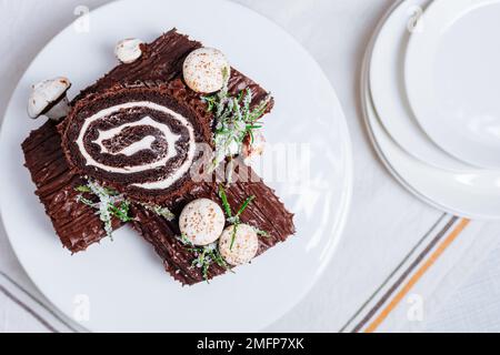 Dessert francese chiamato Yule log o bûche de Noël con funghi merengue e foglie in cima alla vetrata al cioccolato. Posto di fronte all'albero di Natale. Dic Foto Stock