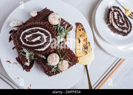 Dessert francese chiamato Yule log o bûche de Noël con funghi merengue e foglie in cima alla vetrata al cioccolato. Posto di fronte all'albero di Natale. Dic Foto Stock