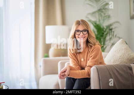 Ritratto di bella donna di mezza età che si rilassa in una poltrona a casa. Donna dai capelli biondi che indossa occhiali e maglione. Foto Stock