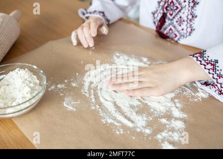 Una ragazza in una maglia nazionale Ucraina ricamata versa la farina sul tavolo per preparare l'impasto Foto Stock