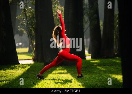 Donna con abito da yoga di colore rosso, che posa i movimenti del guerriero di yoga su uno sfondo verde grasss Foto Stock