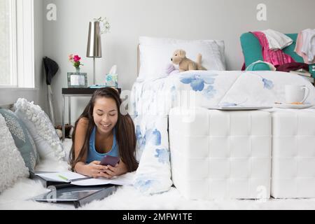 Una ragazza adolescente usa il suo telefono mentre fa il suo lavoro sul pavimento della sua camera da letto. Foto Stock