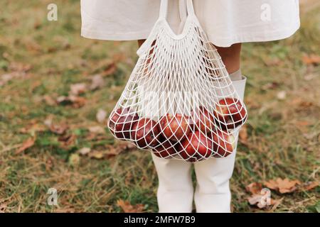 Borsa a corda con mele rosse. Vista delle gambe femminili in stivali bianchi che attraversano il giardino autunnale con borsa ecologica in mano. Concetto di zero sprechi. Nessuna plastica Foto Stock