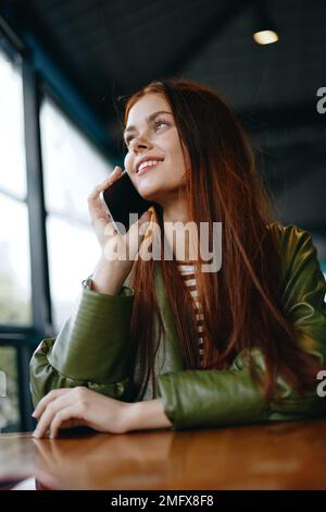 Donna con i capelli rossi blogger freelance seduto in un caffè che parla al telefono e sorridente, ragazza hipster in stile di vita di abiti alla moda Foto Stock