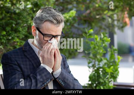 Uomo d'affari maturo che lavora all'aperto con un computer portatile, un uomo anziano che starnutisce dalle allergie, seduto sulla panca del parco nelle giornate di sole, capo in tuta da lavoro. Foto Stock