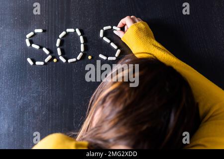 Vista dall'alto di donne depresse non riconoscibili con capelli scuri sdraiati sul tavolo e la disposizione pillole in segnale SOS a casa Foto Stock