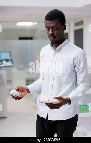 Giovane uomo afroamericano che decide tra due pacchetti di supplementi cardiovascolari in farmacia, tenendo farmaci cuore, leggere istruzioni. Compratore che pensa, trattamento di prescrizione d'acquisto Foto Stock