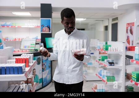 Giovane uomo afroamericano scelta del farmaco di cuore in farmacia, leggendo le istruzioni sui supplementi nutrizionali cardiovascolari. Cliente acquistare farmaci, trattamento su prescrizione medica Foto Stock