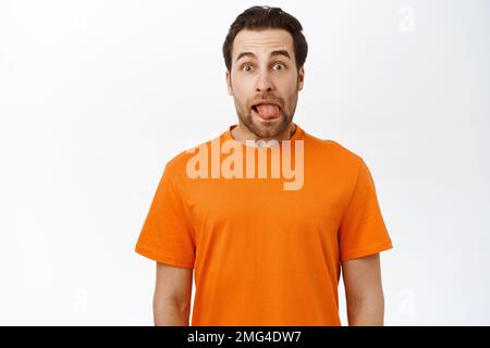 Divertente uomo adulto con barba, mostra la lingua e fa facce sciocco, si erge su sfondo bianco Foto Stock