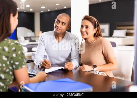 Giovane felice coppia ispanica che consulta con il venditore di mobili quando sceglie il nuovo materasso in negozio Foto Stock