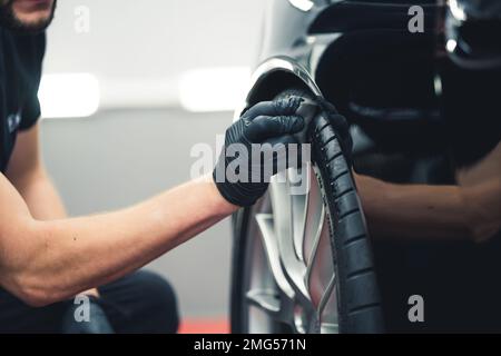 Uomo caucasico che indossa guanti neri stucco pneumatico auto annerimento utilizzando spugna. Dettaglio auto in garage professionale. Primo piano orizzontale in interni. Foto di alta qualità Foto Stock