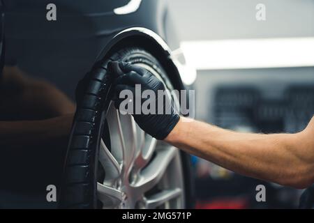 Primo piano di un uomo irriconoscibile che indossa un guanto nero che annerisce le gomme dell'automobile usando la spugna. Auto professionale dettaglio in un garage. Scatto orizzontale in interni. Foto di alta qualità Foto Stock