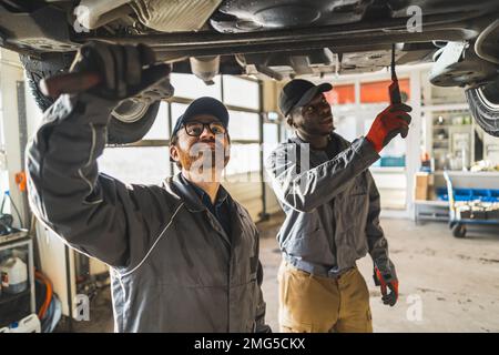 Immagine media di due meccanici che lavorano sul telaio di un'auto e che parlano l'uno con l'altro. Concetto di officina di riparazione. Foto di alta qualità Foto Stock