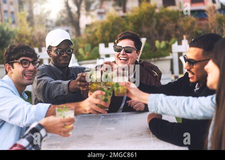 Un gruppo variegato di giovani amici si riunisce intorno ad un tavolo in un pub all'aperto, alzando i bicchieri in un brindisi con mojitos rinfrescante. Festeggia la varietà Foto Stock