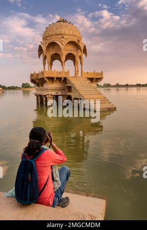 Ragazza indiana viaggiatore, fotografa donna che fotografa Chhatris e santuari con riflesso di loro sulle acque del lago Gadisar. Set da sole e c Foto Stock