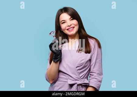 Cosmetician tiene il rullo di pietra per il massaggio del viso. Donna estetista in uniforme e nero guanti in lattice su sfondo blu chiaro studio Foto Stock