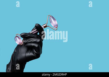 La mano di Beautician può contenere un massaggiatore a rullo cosmetico per il massaggio viso in quarzo rosa. Spazio vuoto. Cosmetologo in lattice nero guanto su dorso blu Foto Stock