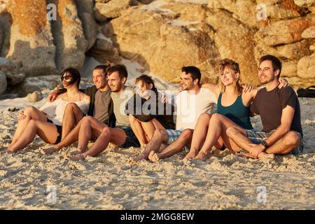 I tramonti sono migliori quando li condividete. un gruppo di giovani amici felici che ammirano insieme il tramonto mentre si siedono sulla spiaggia. Foto Stock