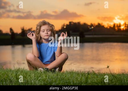 Il bambino pratica yoga e medita in posizione loto vicino al lago al tramonto. Foto Stock