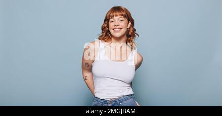Donna con capelli di zenzero sorridente alla macchina fotografica mentre indossa un canotta e jeans. Ritratto di una giovane donna felice in piedi su uno sfondo blu. Foto Stock