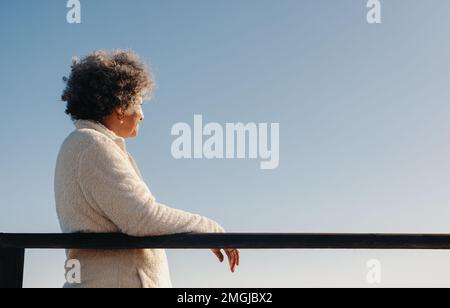 Donna anziana in pensione che guarda una vista dell'oceano mentre sta in piedi su una passerella sulla spiaggia. Felice donna anziana godendo di un rinfrescante holi sul mare Foto Stock