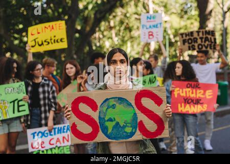 Ragazza musulmana che guarda la macchina fotografica mentre tiene un poster ad un raduno di cambiamento climatico. Gruppo di attivisti giovanili multiculturali che protestano contro la w Foto Stock