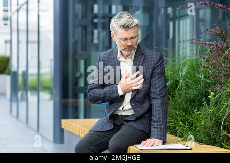 Attacco di cuore in uomo d'affari maturo in aria, capo senior ha dolore al petto grave, l'uomo dai capelli grigi ha dolore al cuore seduto sul banco vicino ufficio. Foto Stock