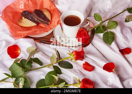 San Valentino colazione a letto tra rose rosse tradizionali - biscotti vegani a forma di cuore e tazza di caffè sulle lenzuola. Estetica, romantico sfondo vacanza lagom. Foto Stock
