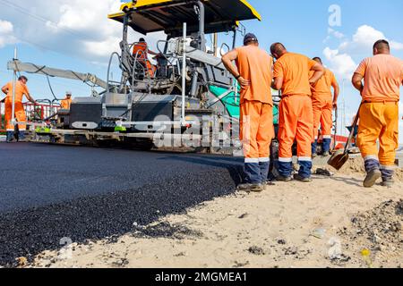 Vista sulla macchina per la posa di asfalto, stendendo strato di asfalto caldo su terreno preparato pochi lavoratori stanno lavorando intorno. Foto Stock