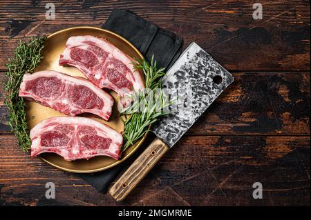 Costolette di lombo di agnello crudo crudo crudo, selle in piatto con rosmarino e timo. Sfondo di legno. Vista dall'alto. Spazio di copia Foto Stock