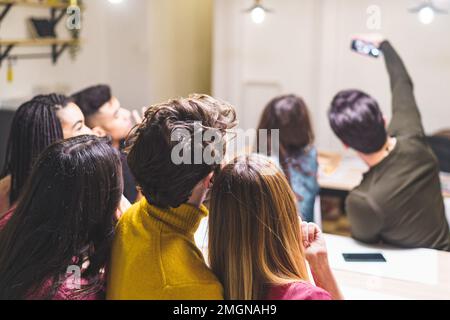 Un gruppo di amici variegati e multiculturali si riunisce in cucina per un rapido selfie. Il colpo insolito cattura il gruppo da dietro - unità e legame o Foto Stock