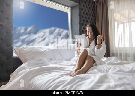 Giovane donna che celebra la vittoria di una lotteria online o ha fatto un buon affare in camera d'albergo con un'espressione allegra del volto. Foto Stock