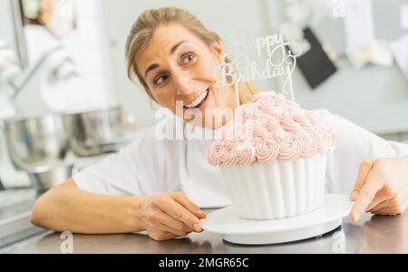 Buon pasticciere sorridente e tenendo grande muffin torta di compleanno con caramelle al forno di pasticceria Foto Stock